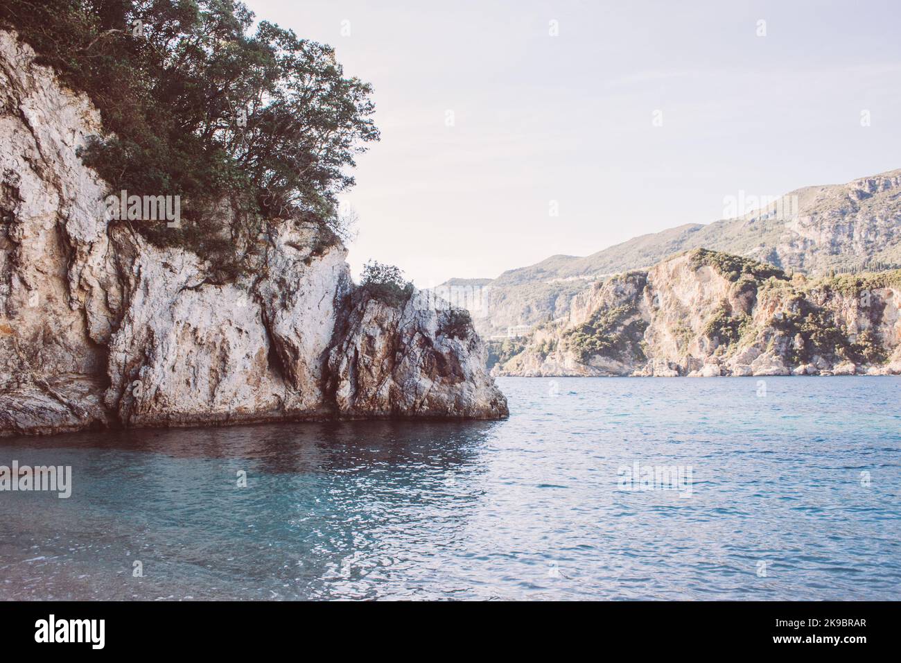 Korfu, Griechenland. Ein riesiger Felsen am Strand mit blauem Wasser, auf dem grüne Pflanzen und Bäume wachsen. Stockfoto