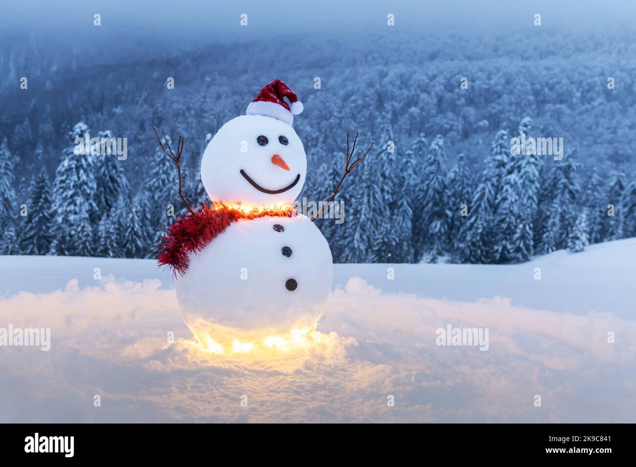 Lustiger Schneemann in stylischem roten Hut und rotem Skalf auf verschneiten Bergen. Leuchtende Girlande für Weihnachtsstimmung Stockfoto