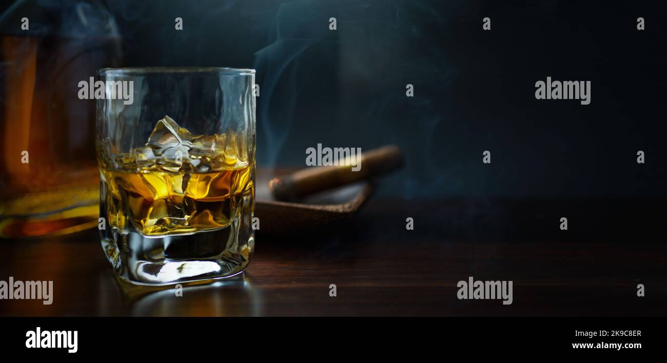 Ein Glas Whiskey mit Eis auf einem Holztisch, eine dampfende kubanische Zigarre und eine Flasche Whiskey in der rauchigen Atmosphäre einer Nachtbar Stockfoto