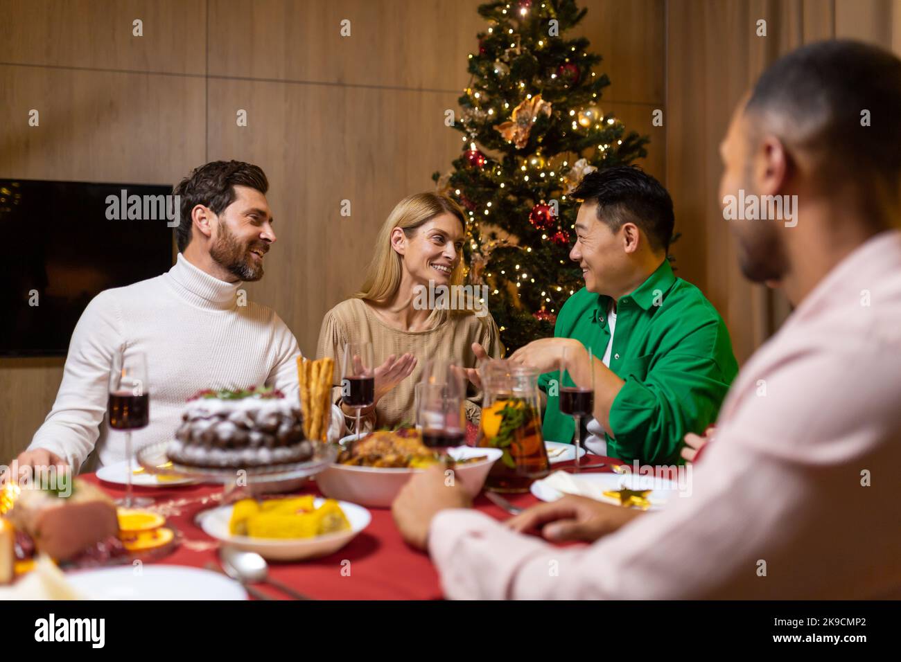 Arbeiten im Unternehmen. Ein Unternehmen aus jungen, schönen, interracial Menschen feiern eine Neujahrsfeier. Sie sitzen am Festtisch neben dem Weihnachtsbaum, reden, lachen. Stockfoto
