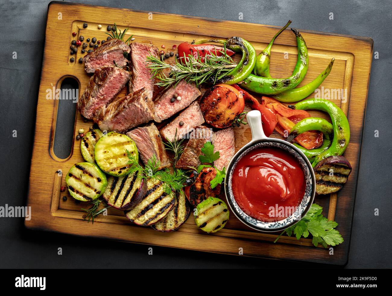 Gebratenes mittelschweres Steak mit gegrilltem Gemüse und Ketchup-Sauce auf Holzschneidebrett auf dunklem Hintergrund Stockfoto