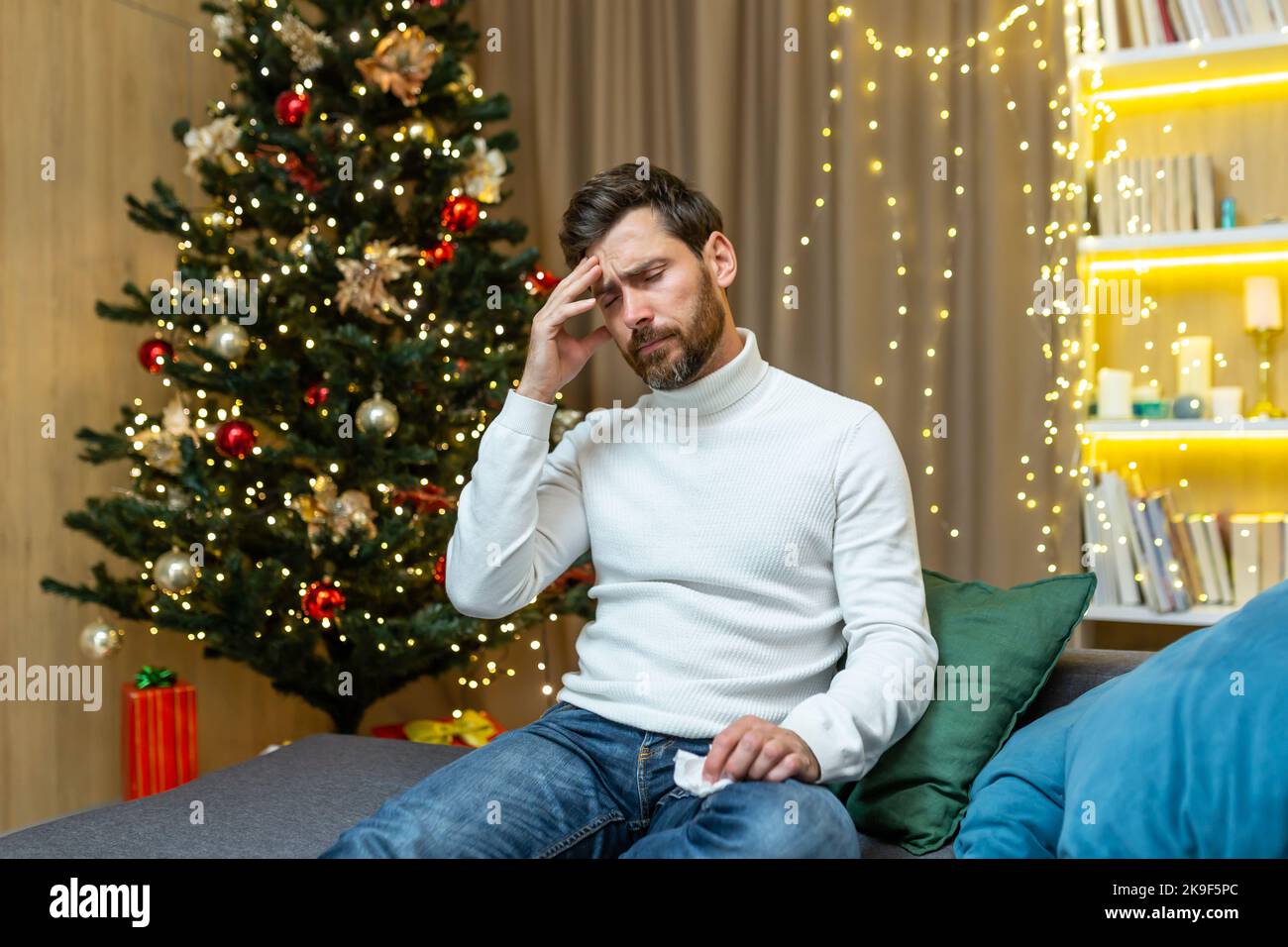 Ein kranker Mann mit Erkältung sitzt zu Weihnachten auf dem Sofa, feiert Neujahr und Weihnachtsferien, hält seinen Kopf mit einer Serviette in den Händen. Stockfoto
