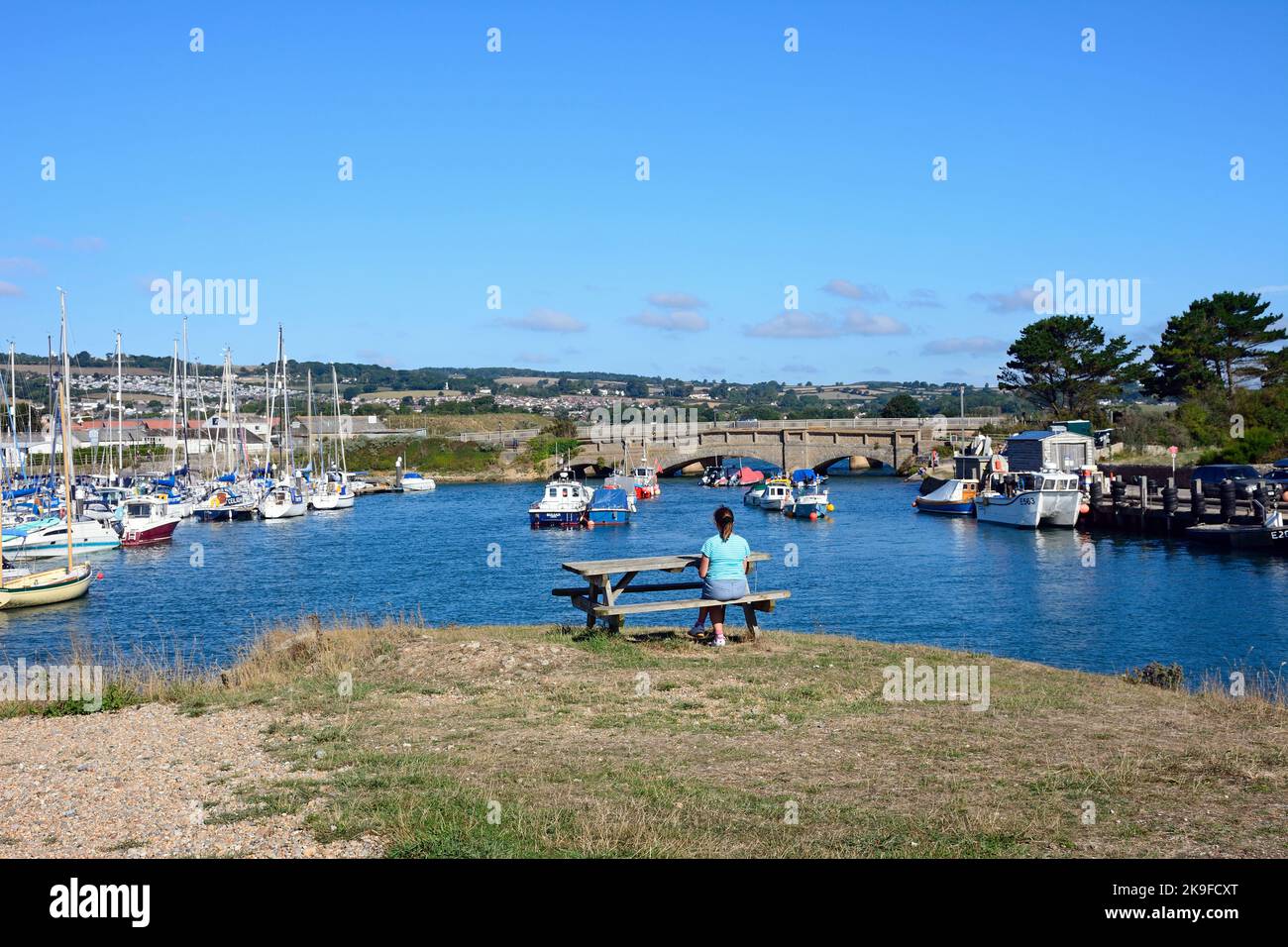 Brücke über die Axe im Hafen und eine Frau, die auf einer Picknickbank sitzt, Axmouth, Devon, Großbritannien. Stockfoto