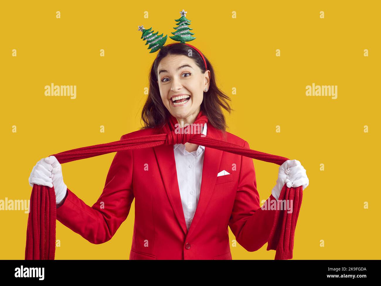 Fröhliche Frau in rotem Anzug und lustiges Weihnachtsband lächelt und bindet roten Schal um den Hals Stockfoto