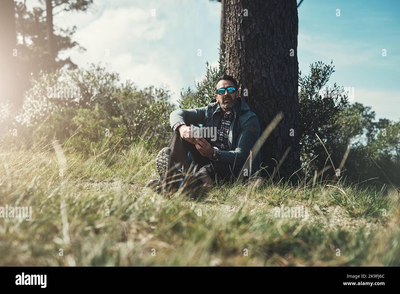 Ich denke, dieser Ort hat die beste Verbindung hier draußen. Porträt eines Mannes mittleren Alters, der an einem Baum sitzt und sein Handy benutzt. Stockfoto