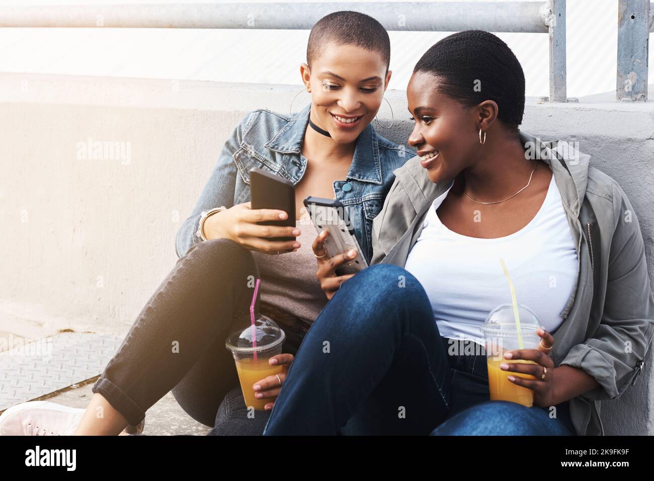 Zwei junge Frauen in der Stadt sitzen sich, während sie lachen und ihre Getränke durch Textnachrichten lesen. Stockfoto