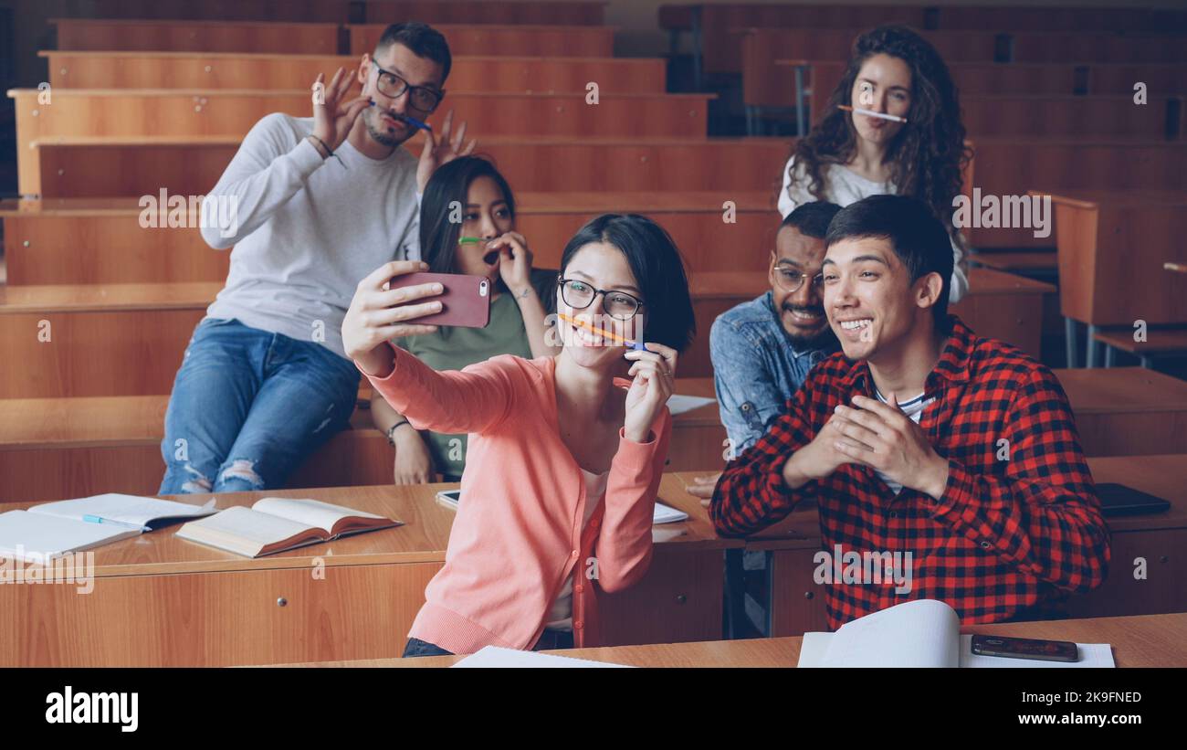 Spielerische junge Leute Studenten nehmen lustige Selbsthilfe mit Stiften und Bleistiften als Schnurrbart, posieren und zeigen Handgesten Daumen-nach oben und V-Zeichen sitzen im Klassenzimmer. Stockfoto