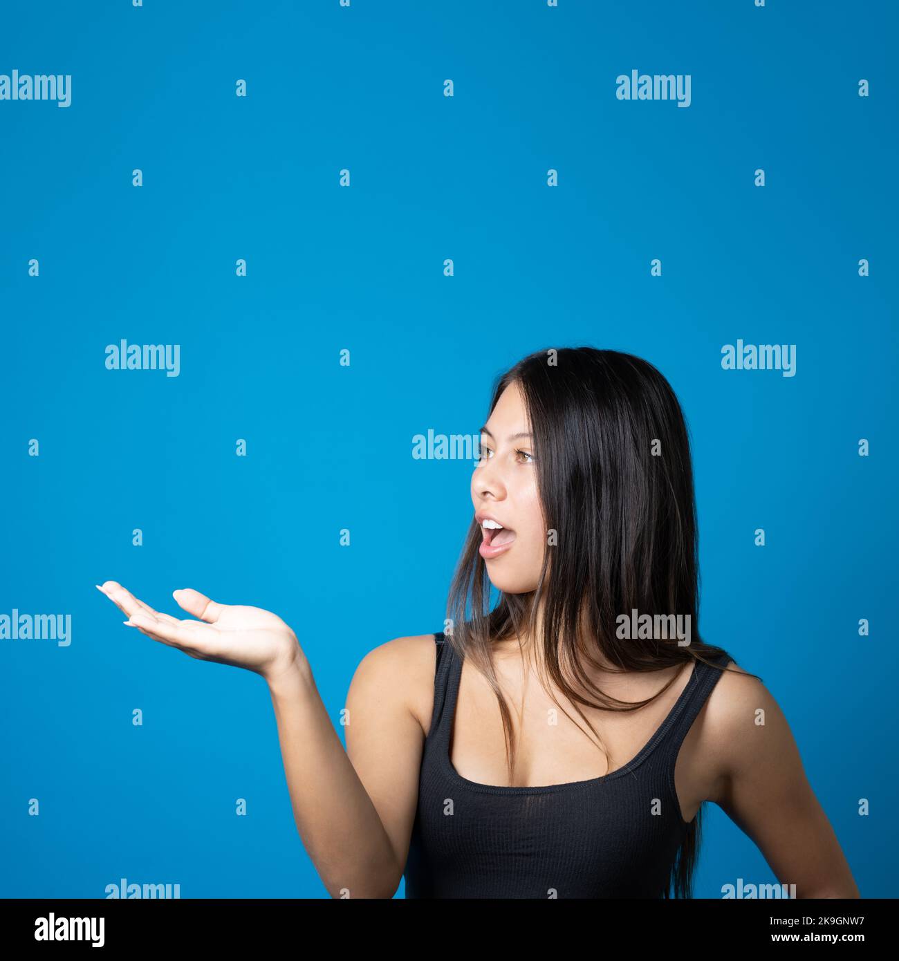 Junge trendige schockierte Frau, die Produkt oder Text ansieht und Kopierraum auf ihrer Handfläche hält und präsentiert, isoliert auf blauem Rücken Stockfoto