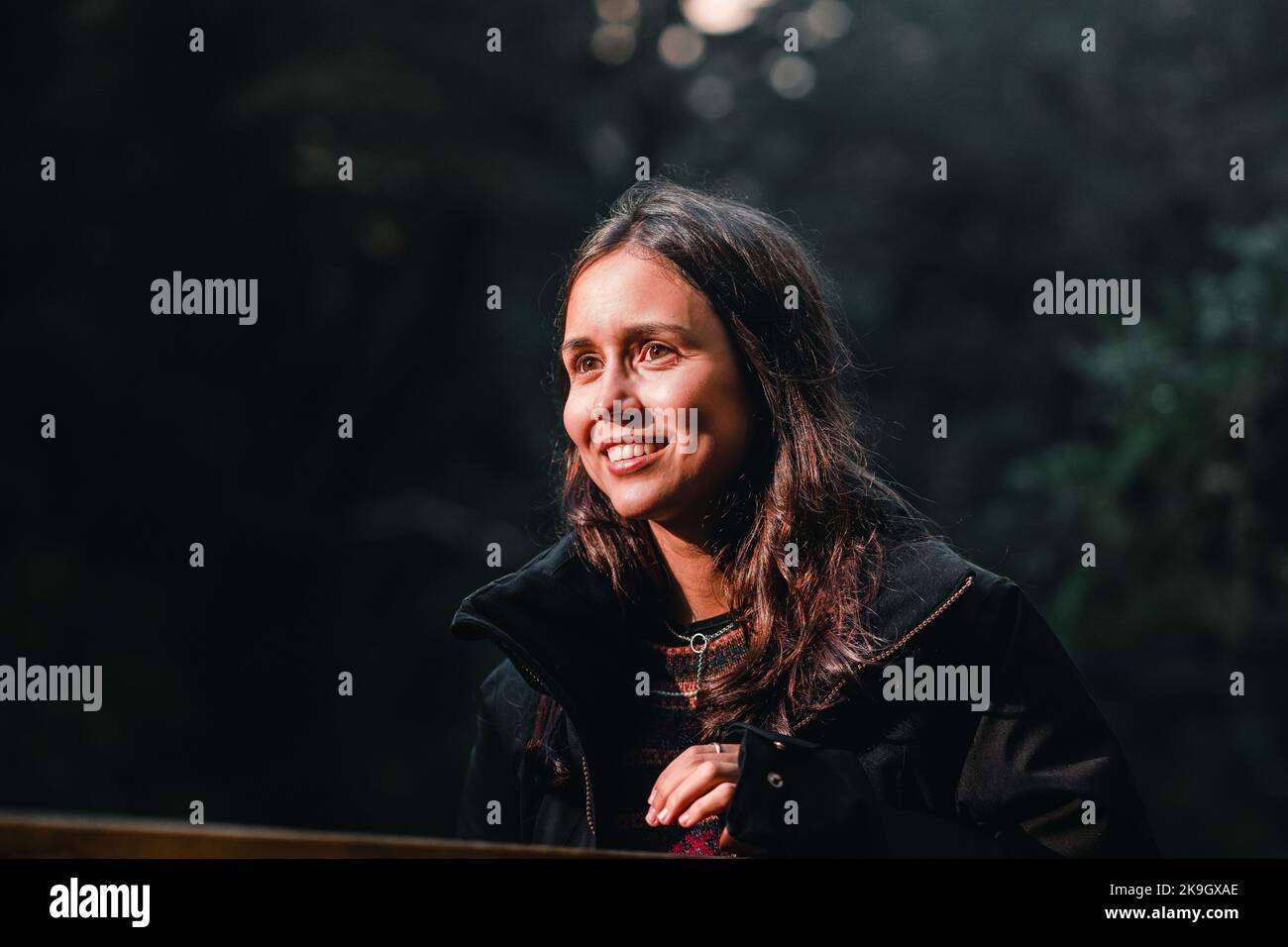 Junge kaukasische Brünette mit einem weißen Lächeln, gekleidet in einen Wollpullover und eine schwarze Jacke mit einer Halskette um ihren Hals mit der Sonneneinstrahlung Stockfoto