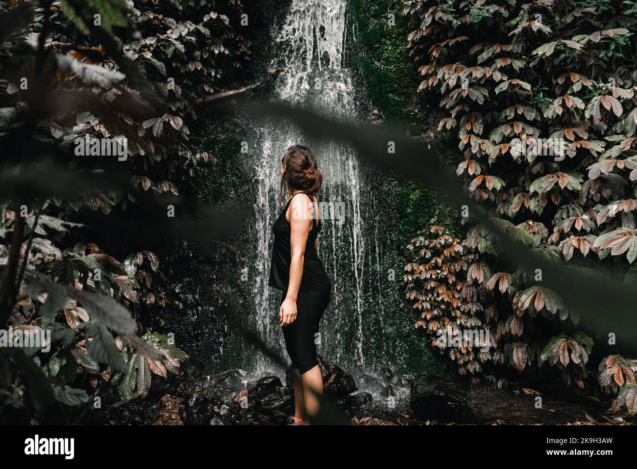 kaukasisches Mädchen mit gesammeltem braunen Haar in schwarzer Kleidung, das den Wasserfall zwischen Bäumen und Waldvegetation betrachtet Stockfoto