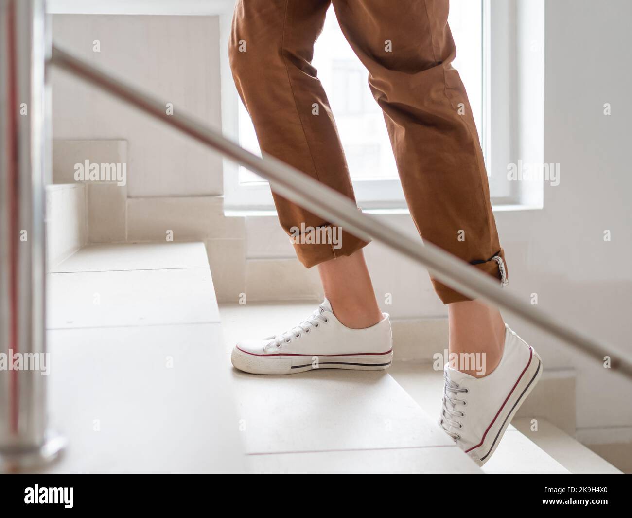 Frau in weißen Turnschuhen und khakifarbenen Hosen geht nach oben in ihre Wohnung. Weiße Treppe im Mehrfamilienhaus. Lässiges Outfit, urbane Mode. Stockfoto