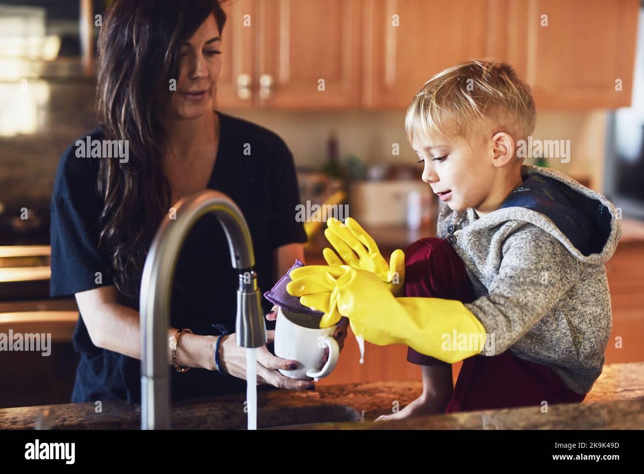 Mein kleiner Helfer. Ein fröhlicher kleiner Junge trägt gelbe Waschhandschuhe und hilft seiner Mutter, in der Küche zu Hause zu kochen. Stockfoto