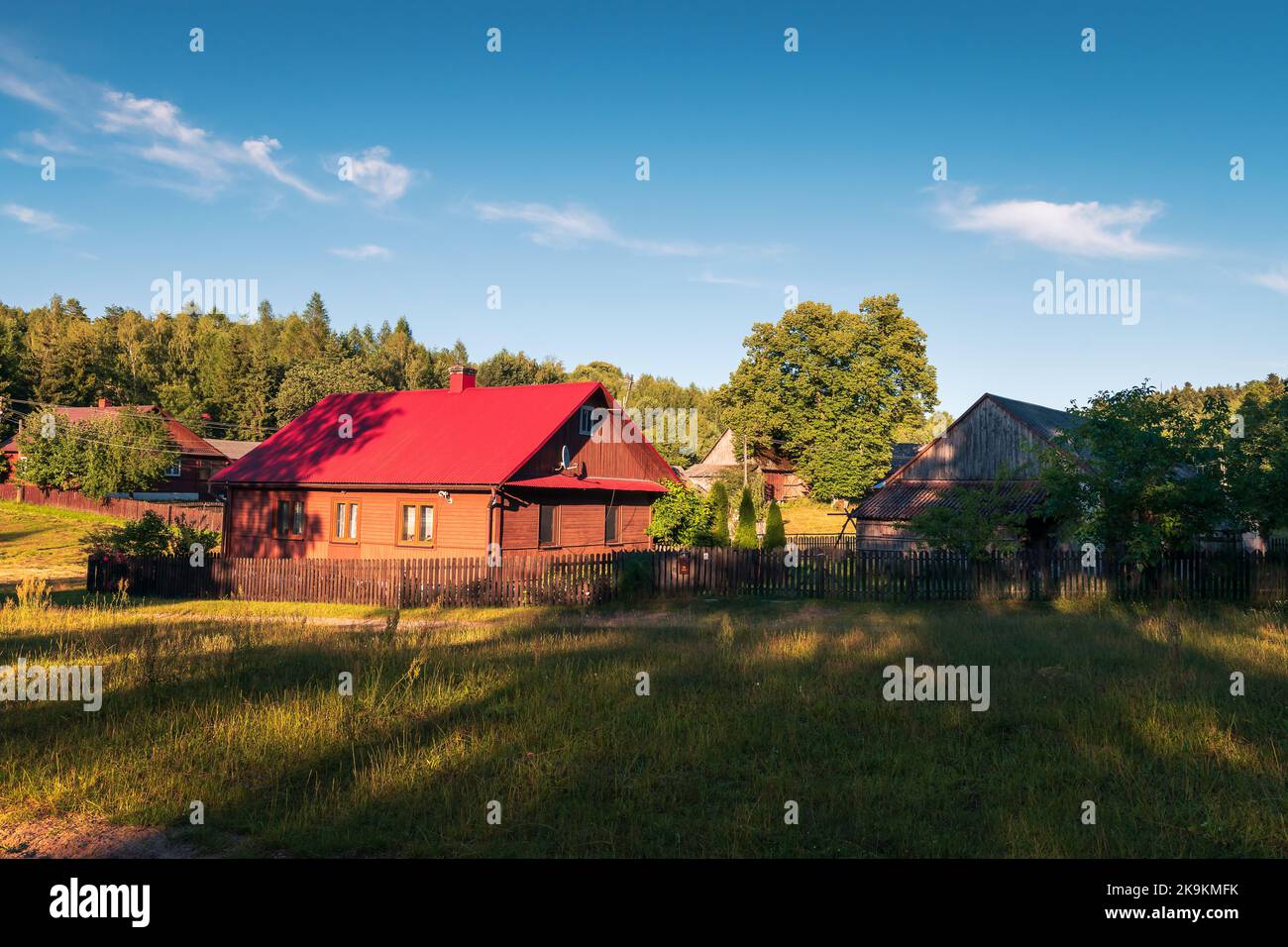 Schöne ländliche Siedlung im Licht der Nachmittagssonne. Ein Haus mit einem roten Dach und einem Holzzaun im Vordergrund, Krasnobród, Roztocze Stockfoto