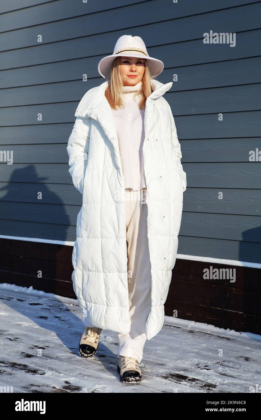 Junge Frau posiert in totaler weißer Mode Sport chic Look mit langer Daunenjacke, Wolle Kostüm und Fedora. Modisches blondes Modell, winterlicher Street-Stil Stockfoto
