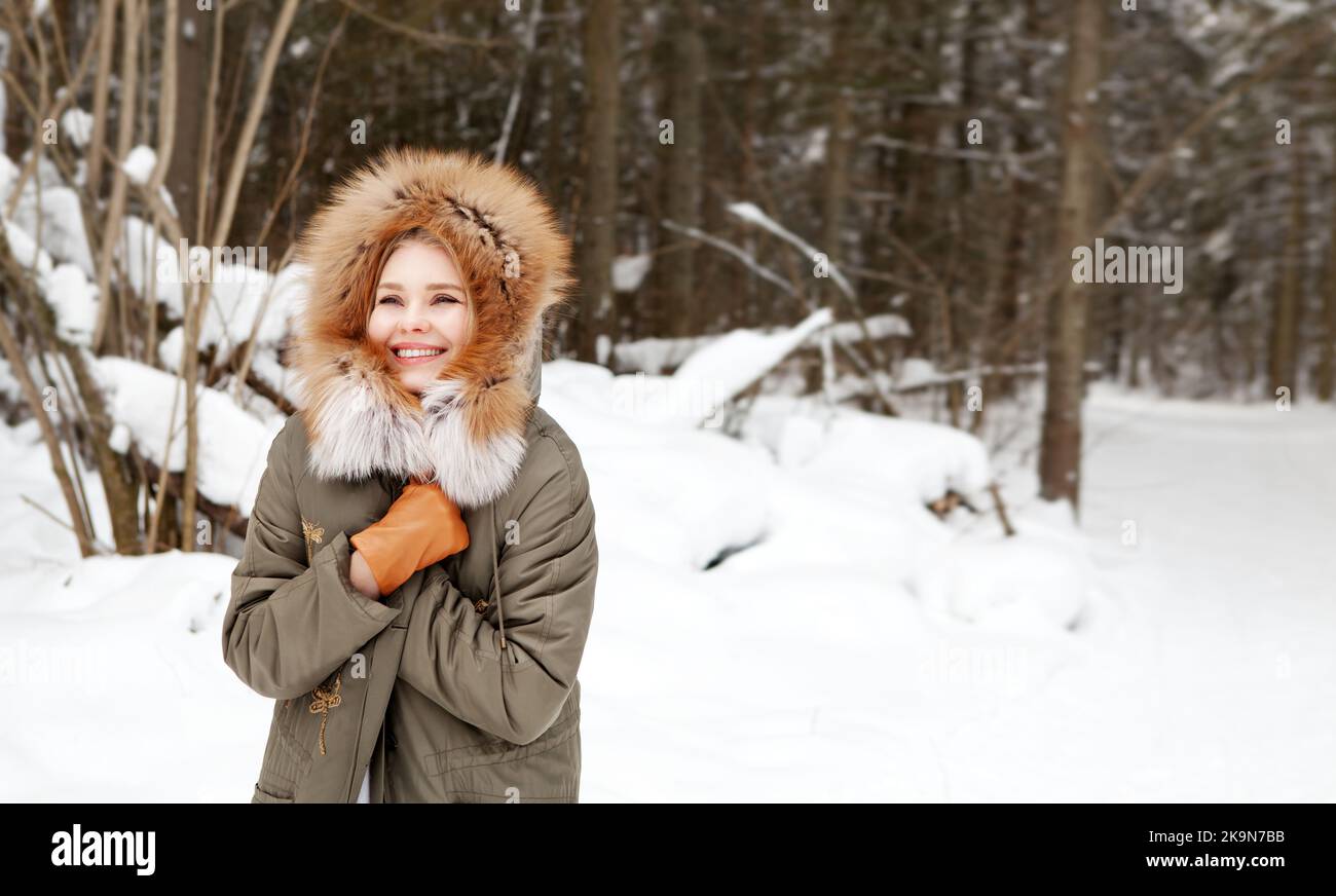 Glückliche Frau lächelt und hüllt sich in warme Kleidung im Winterwald auf Schnee Hintergrund. Tragen stilvolle Mode-Outfit khaki Parka Mantel Jacke mit Stockfoto