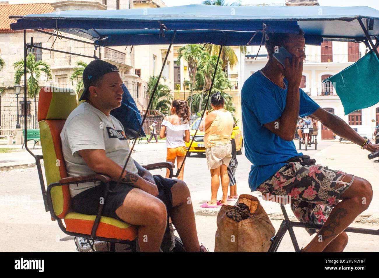Ein junger Mann trägt einen Beutel mit Ananas in einem Pediküre oder Bicitaxi. Der Fahrer des Fahrzeugs spricht auf einem Smartphone. Stockfoto