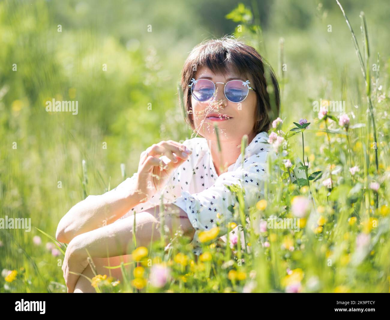Frau in bunten Sonnenbrillen, genießt Sonnenlicht und Blumenduft auf Grasfeld. Sommerliche Stimmung. Entspannen Sie sich im Freien. Selbstberuhigend. Stockfoto