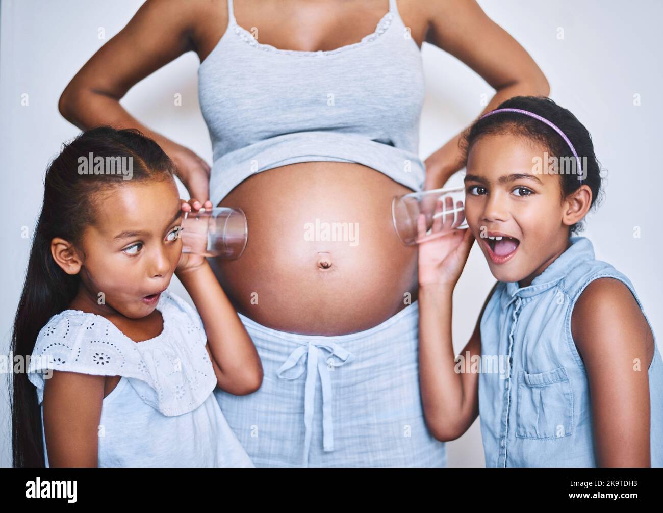 Ich habe etwas gehört. Portrait von zwei fröhlichen kleinen Mädchen, die neben ihrer Mutter stehen, während sie jeweils ein Glas auf ihren schwanger Bauch legen, um zuzuhören Stockfoto