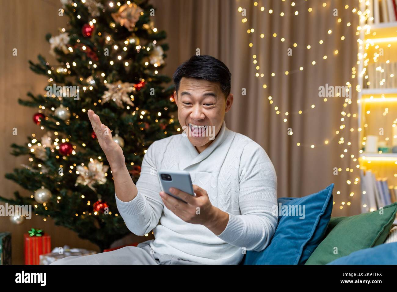 Weihnachtsmann in der Nähe von Baum zu Hause lesen gute Nachrichten aus dem Telefon bekam Gewinn und Rabatt im Online-Casino, asiatische Mann hält Hand nach oben Geste des Triumphs sitzen auf dem Sofa im Wohnzimmer an Neujahr Urlaub. Stockfoto