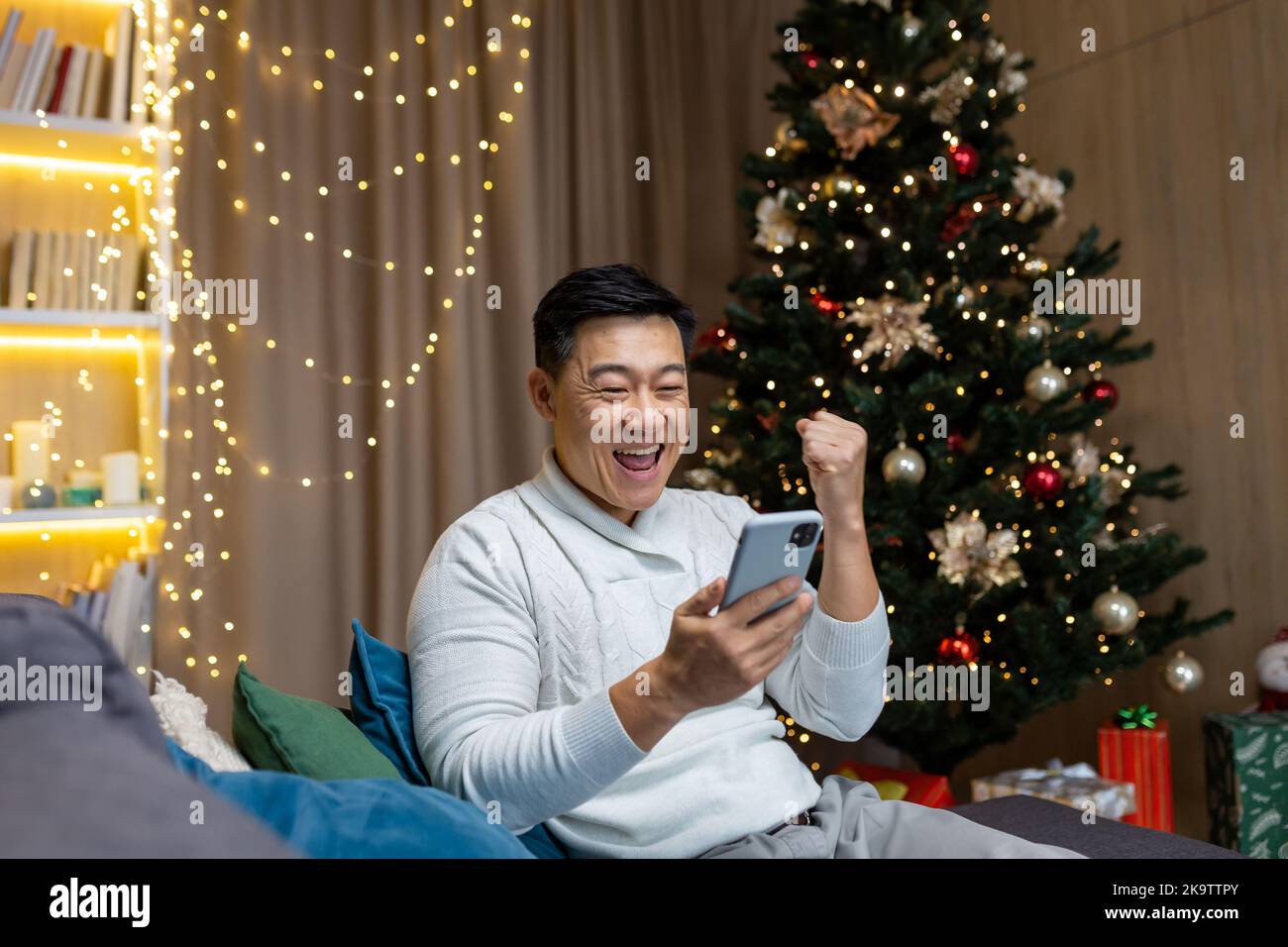 Weihnachtsmann in der Nähe von Baum zu Hause lesen gute Nachrichten aus dem Telefon bekam Gewinn und Rabatt im Online-Casino, asiatische Mann hält Hand nach oben Geste des Triumphs sitzen auf dem Sofa im Wohnzimmer an Neujahr Urlaub. Stockfoto