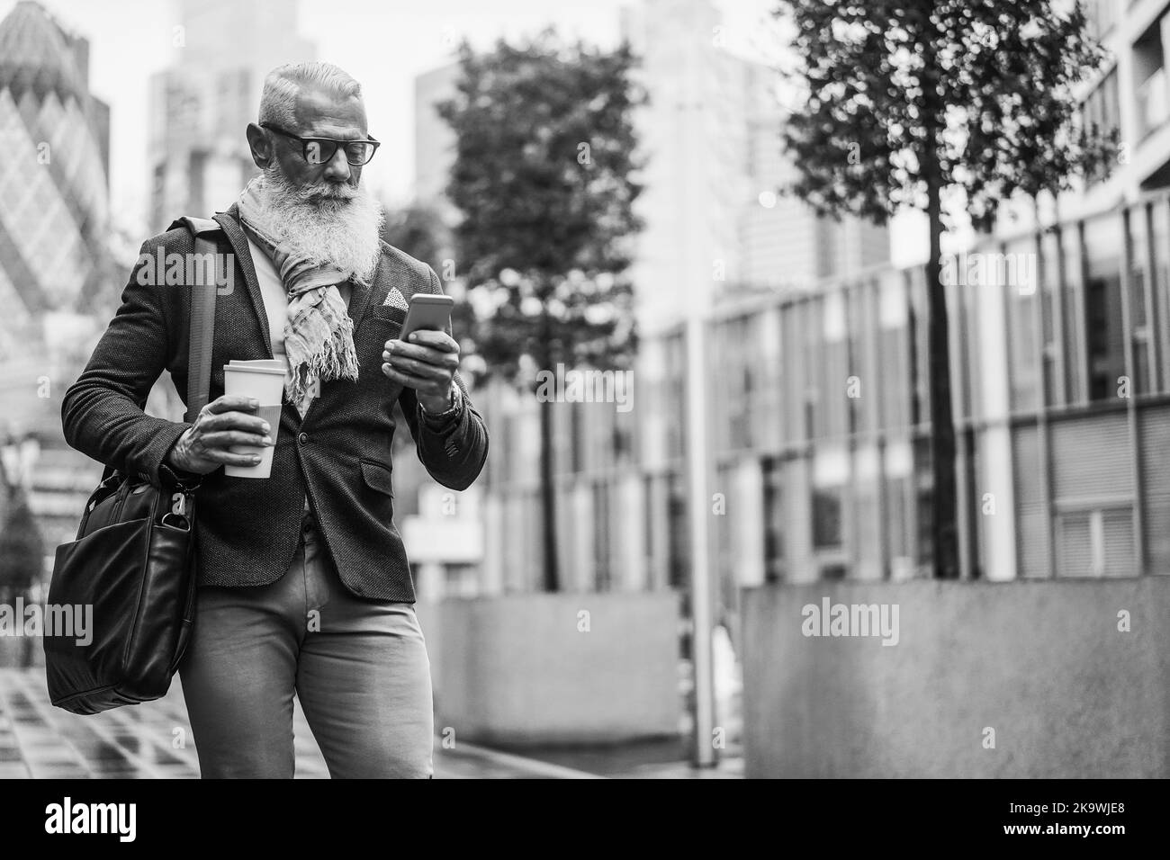 Business Hipster Senior Mann mit Mobiltelefon, während zu Fuß mit Stadt im Hintergrund arbeiten - Focus Face - Schwarz-Weiß-Schnitt Stockfoto