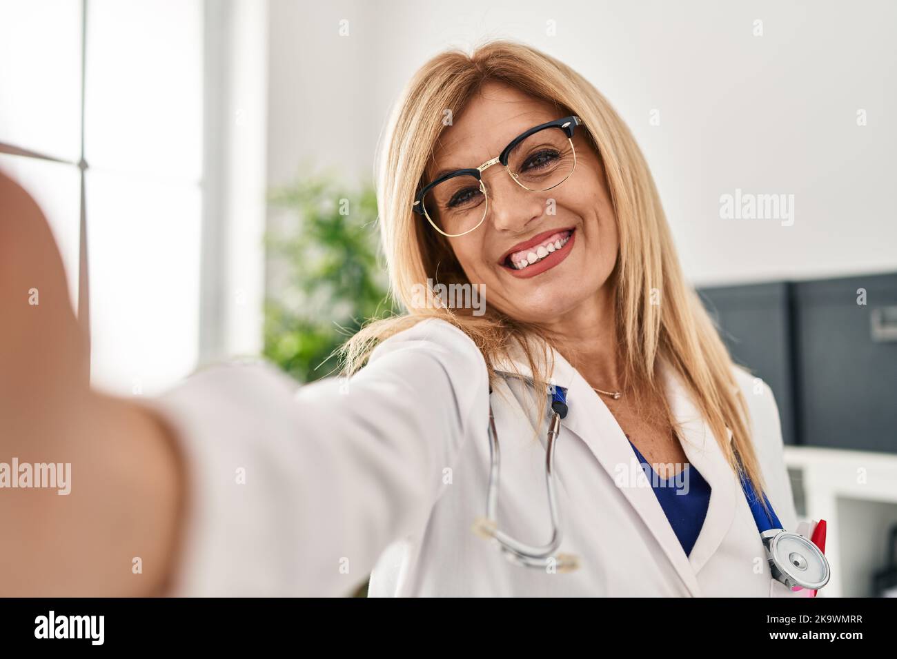 Blonde Frau mittleren Alters in Arztuniform machen Selfie bei der Kamera in der Klinik Stockfoto