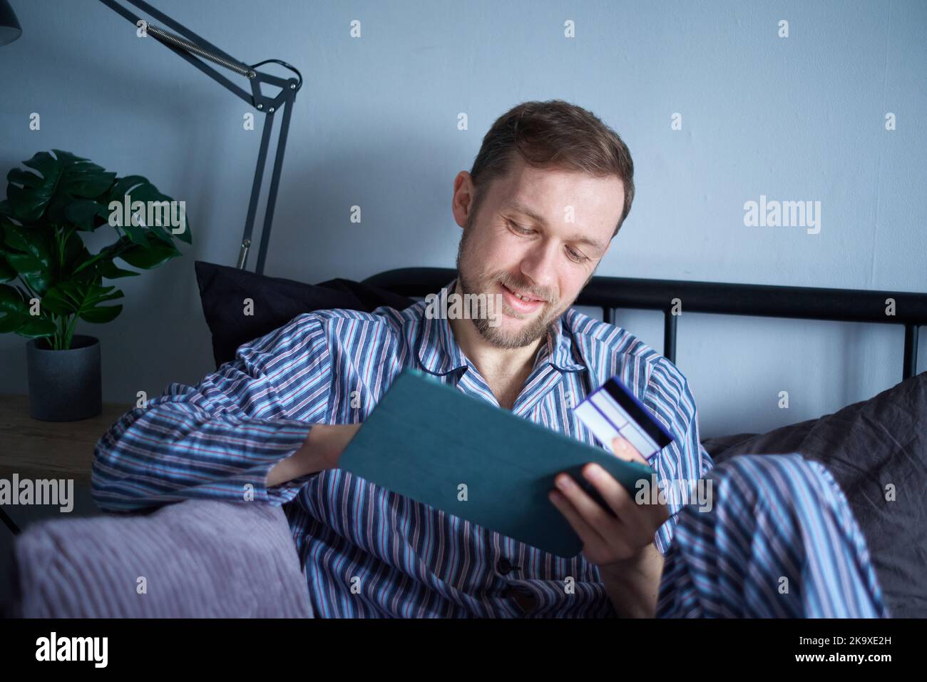 Junge attraktive Mann mit Tablet-Computer im Bett am Morgen sitzen, die Einkäufe online. Kaukasischer Mann mit Touchpad zum Einkaufen mit Kreditkarte in den Händen. Hochwertige Bilder Stockfoto