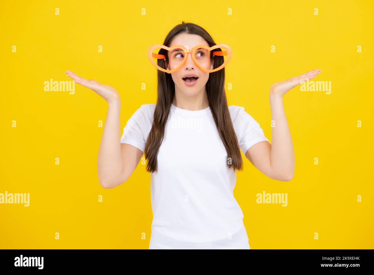 Schönheit lustige Teenager-Mädchen mit Party-Brille, gelber Hintergrund. Überraschte junge Frau mit herzförmigen Gläsern. Stockfoto
