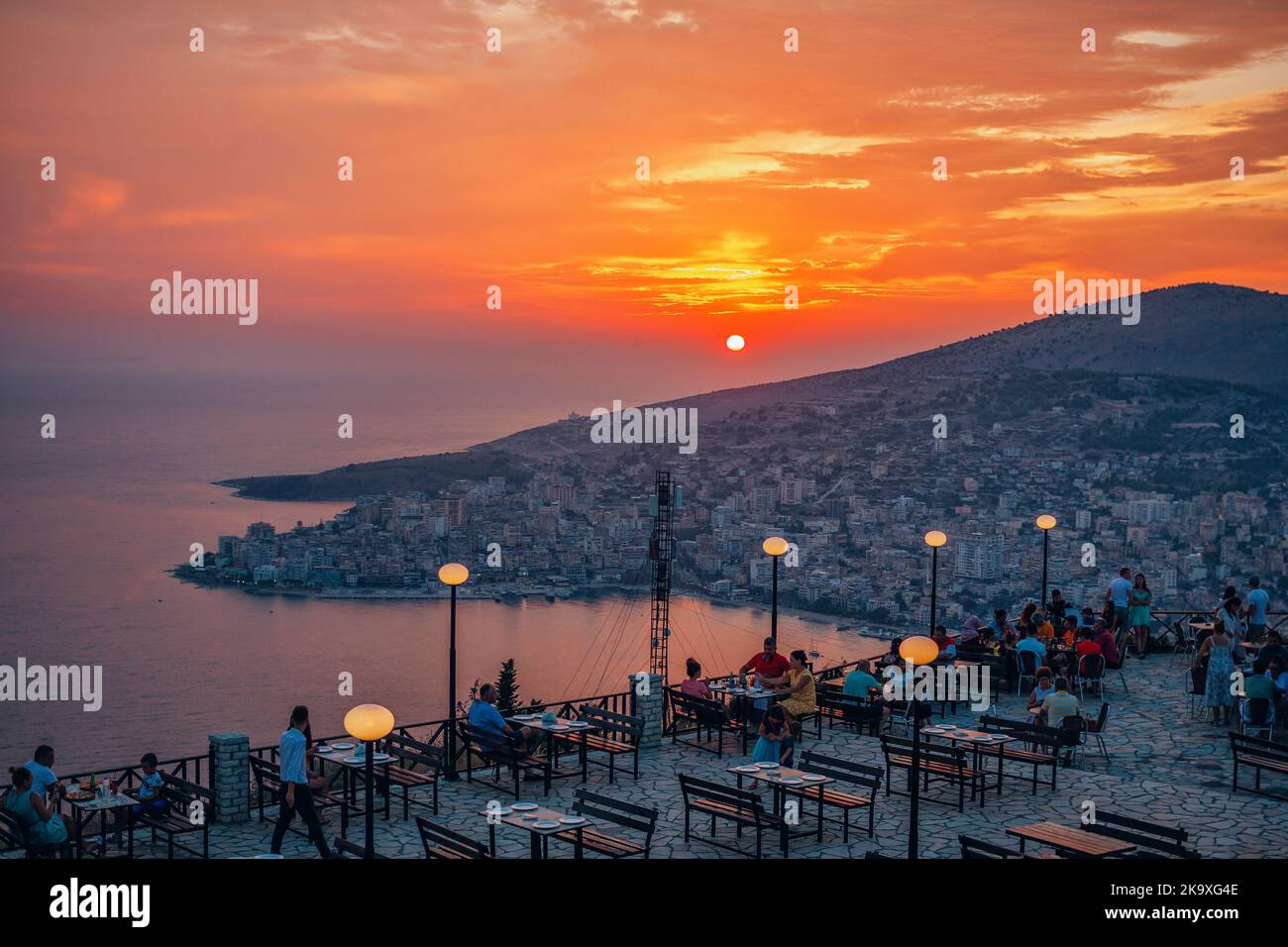 Saranda, Albanien – 1. August 2020: Unvergleichliches Panorama der Stadt bei Sonnenuntergang, Meer, Bergen, farbenfrohem Himmel, Café im Freien und Menschen, die die Aussicht genießen Stockfoto