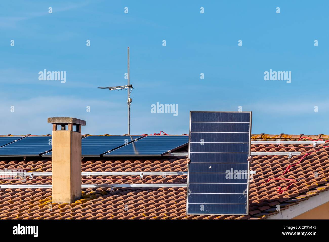 Ein Solarmodul wird von einem Techniker angehoben, der sich darauf vorbereitet, es auf dem Dach eines rot gefliesten Hauses für eine Photovoltaikanlage zu montieren Stockfoto