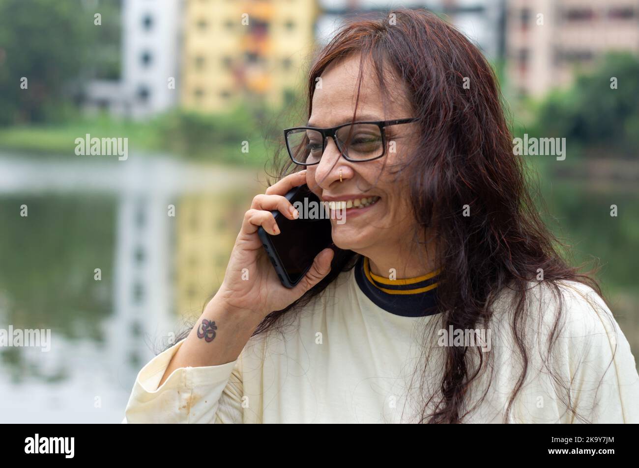 Lächelnde Frau mit langen braunen Haaren bei mittleren Erwachsenen, die im Freien auf dem Mobiltelefon spricht. Nahaufnahme. Sie trägt weißes T-Shirt und Specs. Vorderansicht. Kopf und Stockfoto