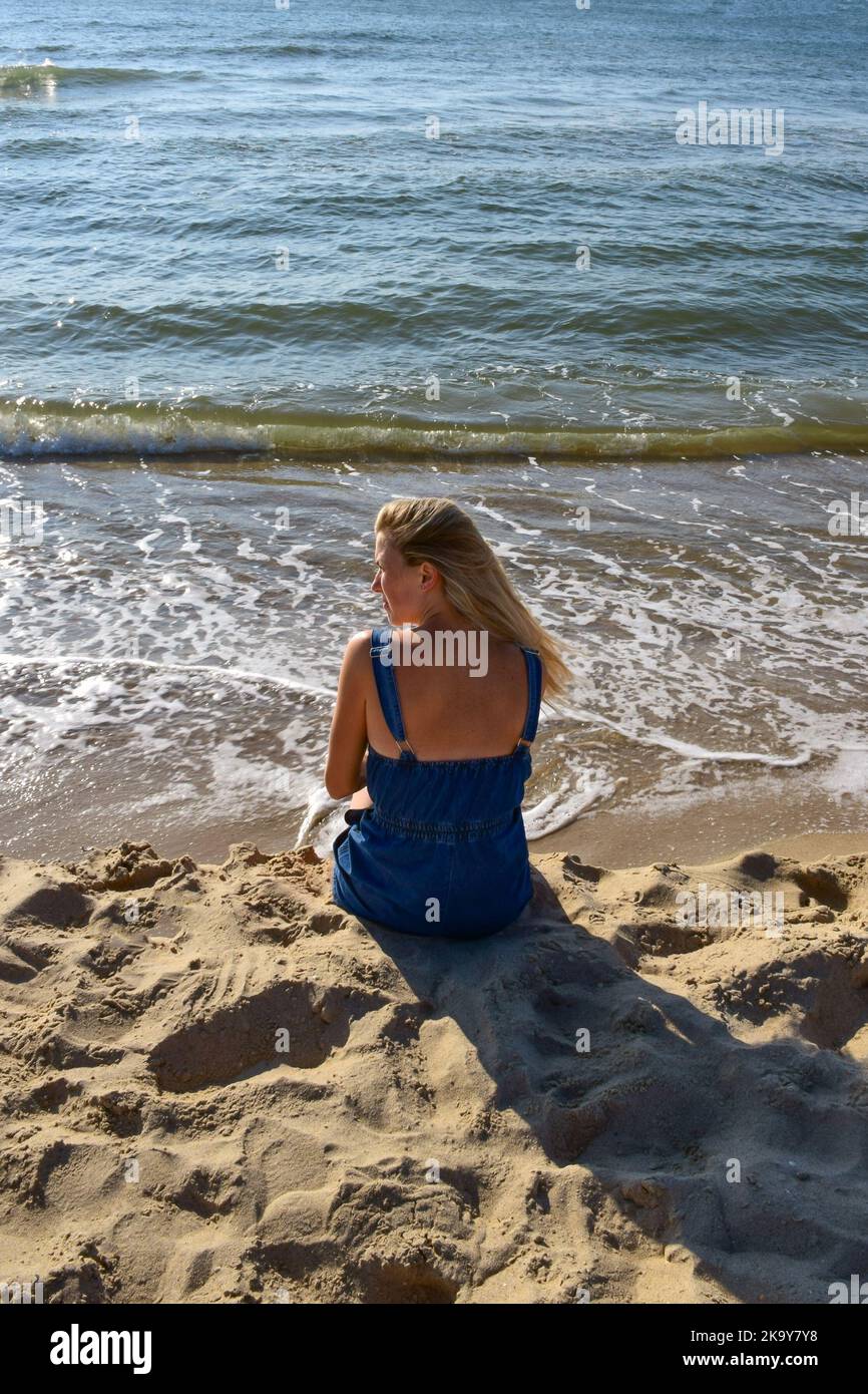 Eine junge Frau sitzt auf dem Sand am Strand in der Nähe des Meeres und blickt in die Ferne. Rückansicht Stockfoto