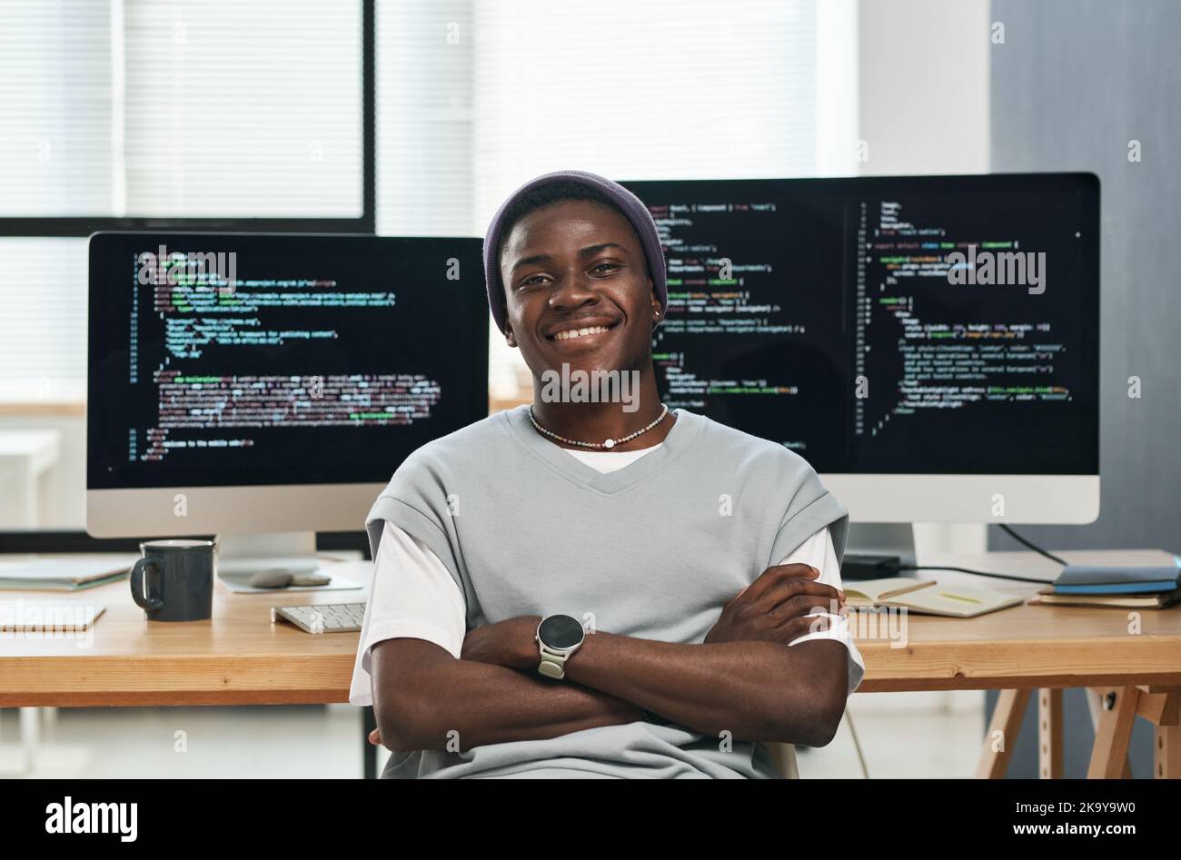 Ein glücklicher junger erfolgreicher IT-Ingenieur im Bereich Casualwear, der Sie beim Sitzen am Arbeitsplatz mit kodierten Daten vor Computerbildschirmen ansieht Stockfoto