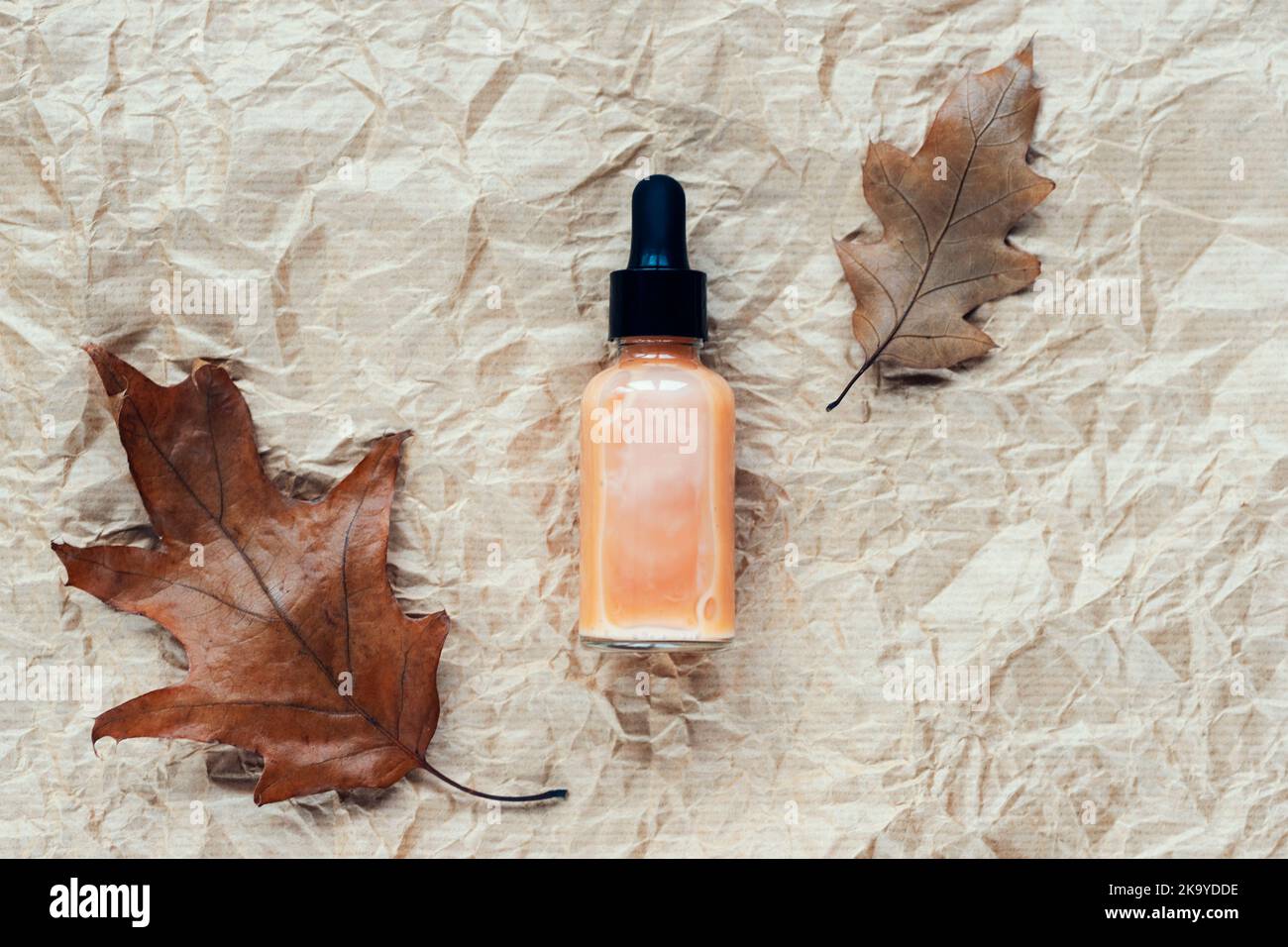 Serum-Tropfflasche und braune Herbstblätter auf beigefarbenem, zerknittertem Papierhintergrund. Draufsicht, flach liegend. Stockfoto
