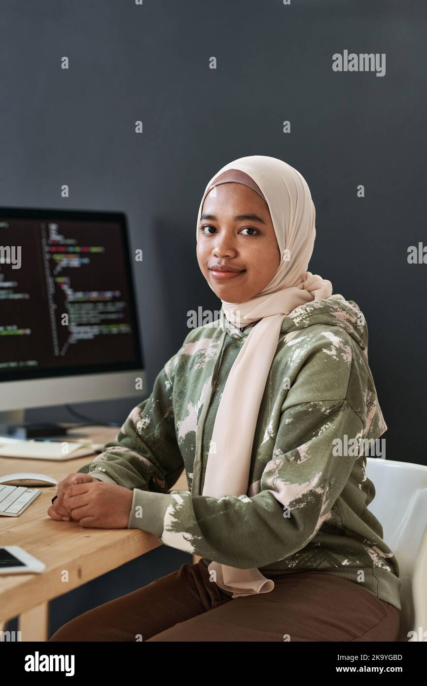 Junge erfolgreiche muslimische Geschäftsfrau in Casualwear, die am Arbeitsplatz mit kodierten Daten gegen den Computerbildschirm sitzt Stockfoto