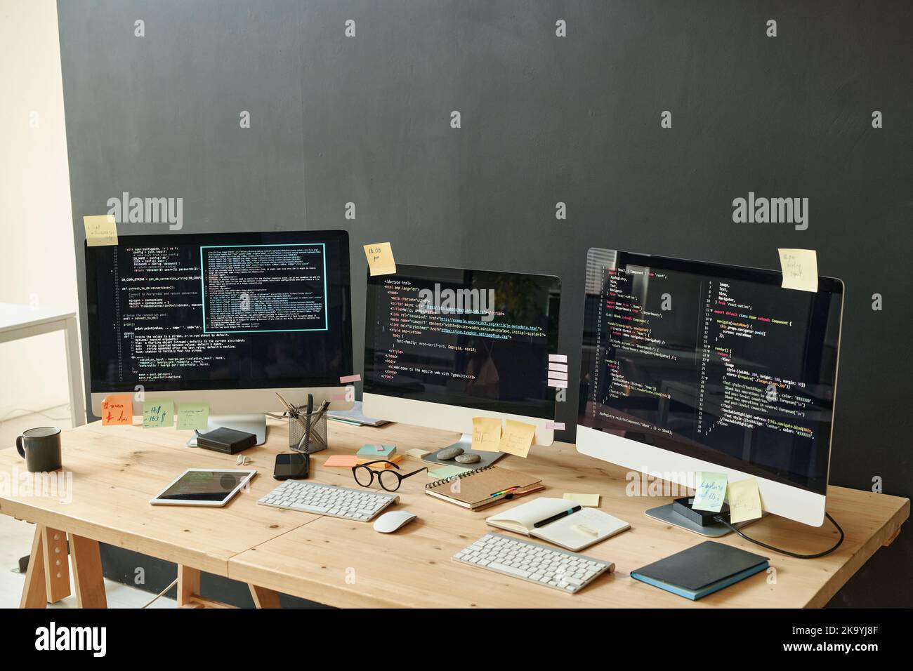 Drei Computermonitore mit kodierten Daten und Haftnotizpapieren auf ihren Bildschirmen, die am Arbeitsplatz eines modernen IT-Ingenieurs im Büro stehen Stockfoto