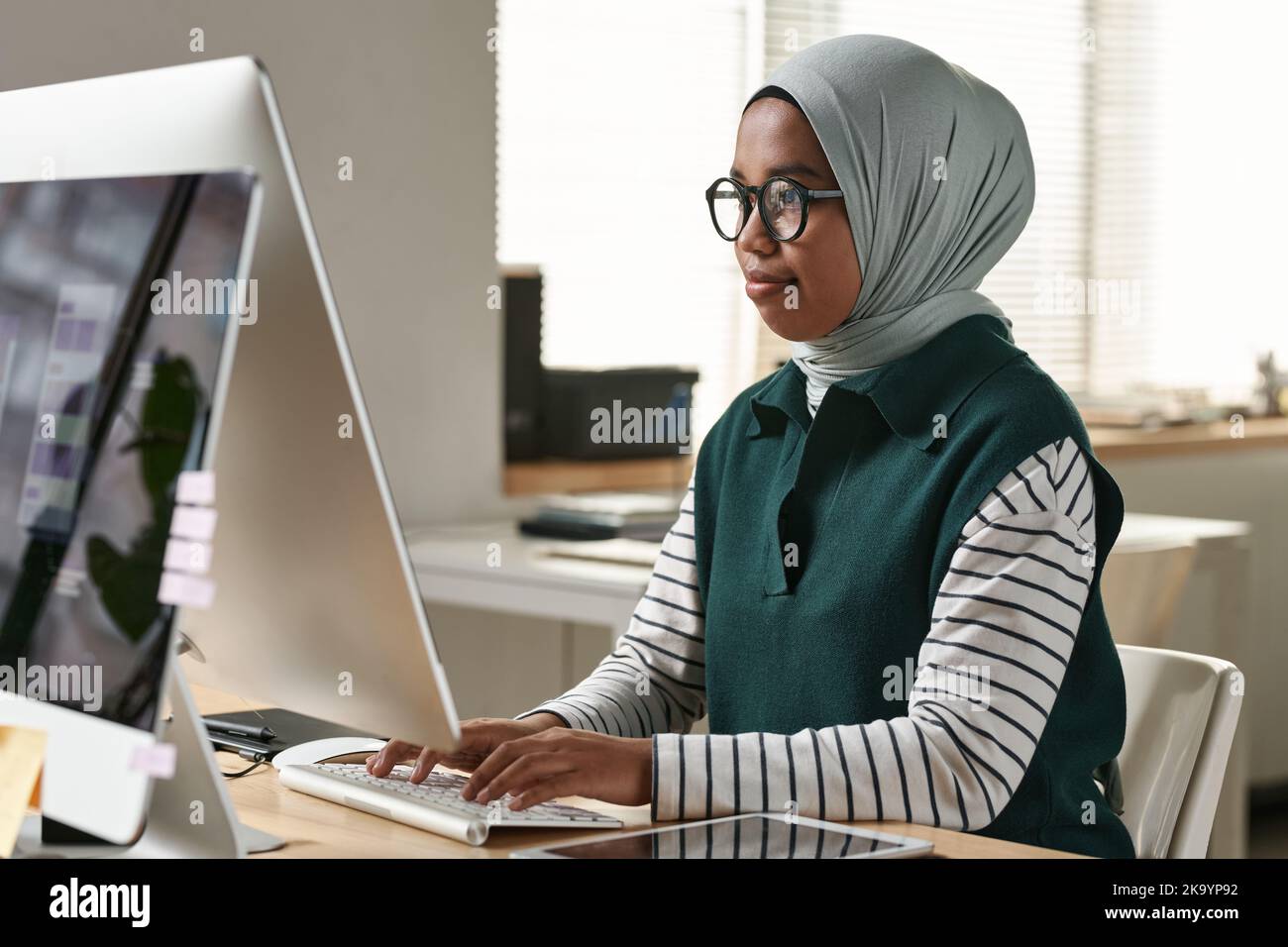 Junge selbstbewusste muslimische Praktikantin oder Designerin in Brillen, Hijab und Casualwear Networking vor dem Computermonitor Stockfoto