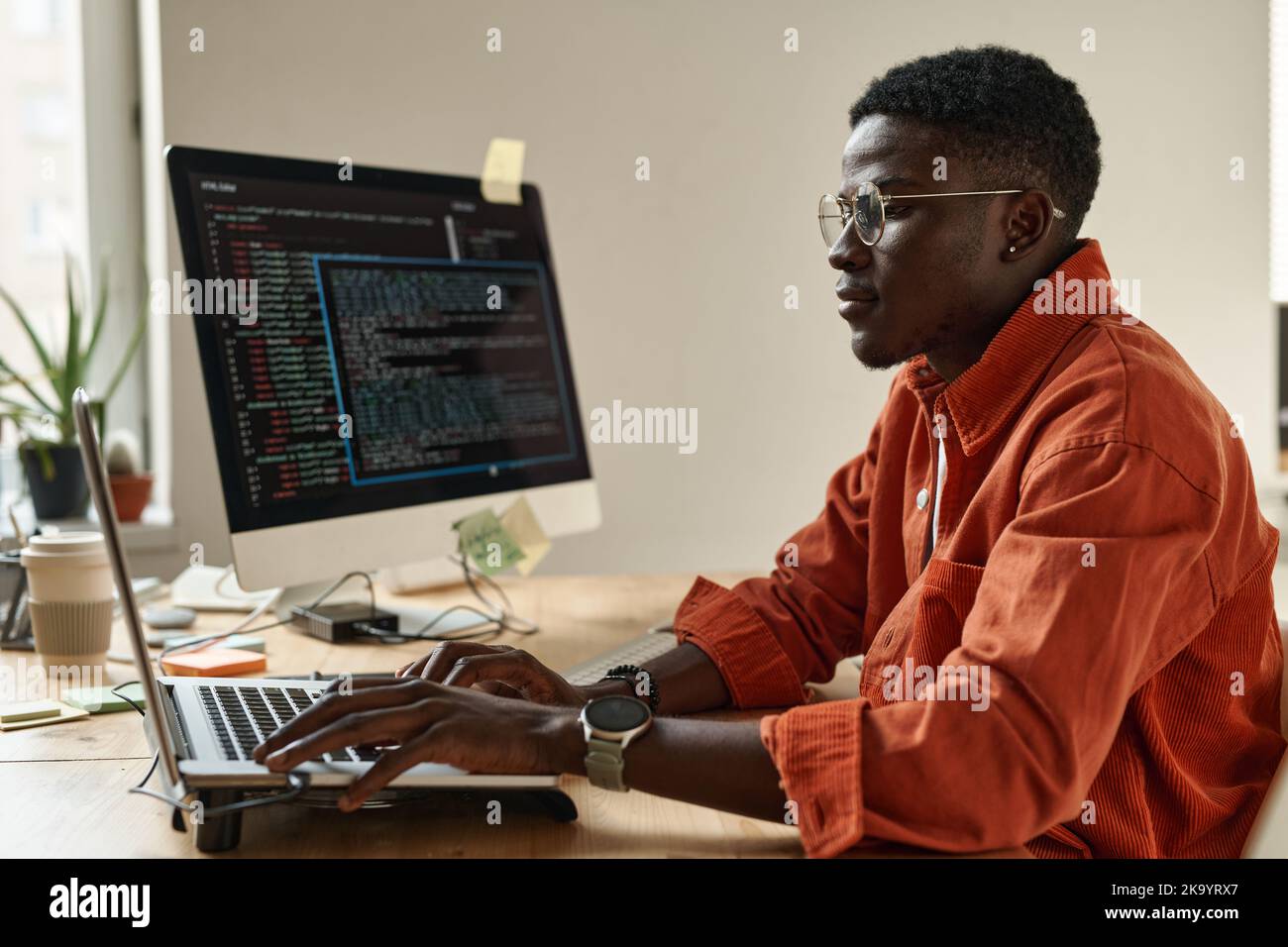 Seitenansicht eines jungen schwarzen Mannes in eleganter Freizeitkleidung und einer Brille, die Daten entschlüsselt, während er vor dem Laptop am Arbeitsplatz sitzt Stockfoto