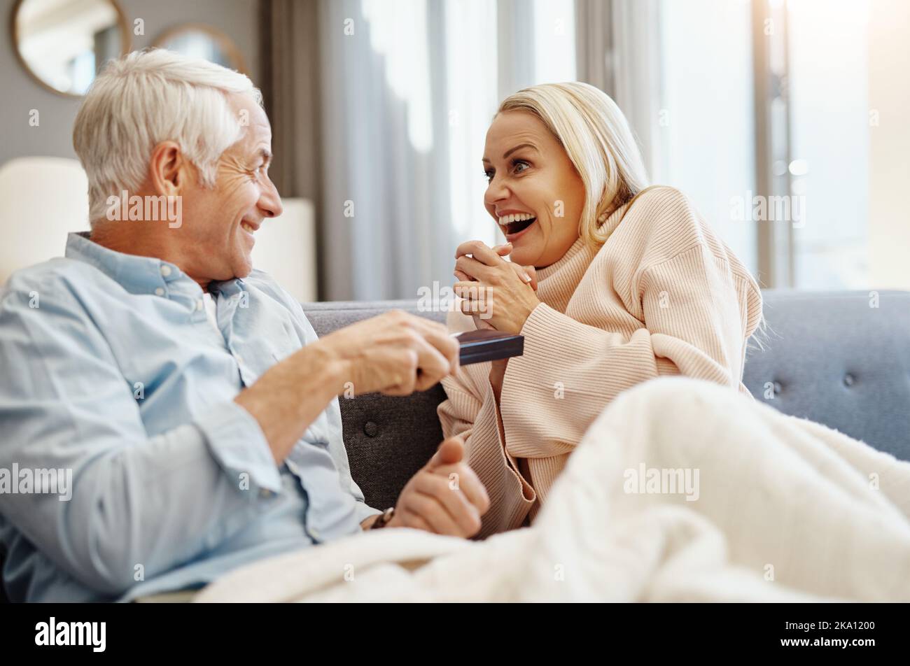 Geschenk geben ist eine der größten Freuden des Lebens. Reifer Mann gibt seiner Frau ein Geschenk auf dem Sofa zu Hause. Stockfoto