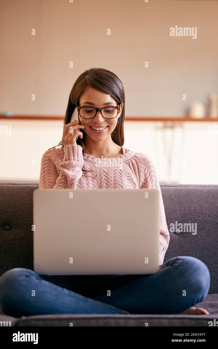 Auf der bequemen Couch hält sie sich auf dem Laufenden. Eine attraktive junge Frau, die zu Hause einen Laptop und ein Telefon auf dem Sofa nutzt. Stockfoto