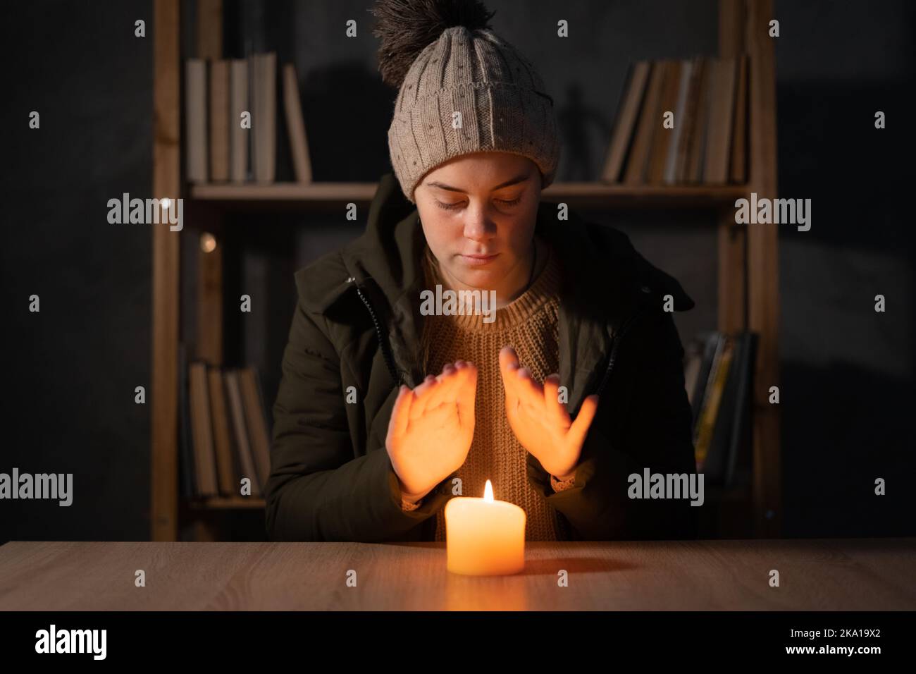 Frierende junge Frau in Winterkleidung wärmt ihre Hände auf Lichter mit Kerzen. Leidet in keiner Heizung und keinen Strom während einer Energiekrise in Stockfoto