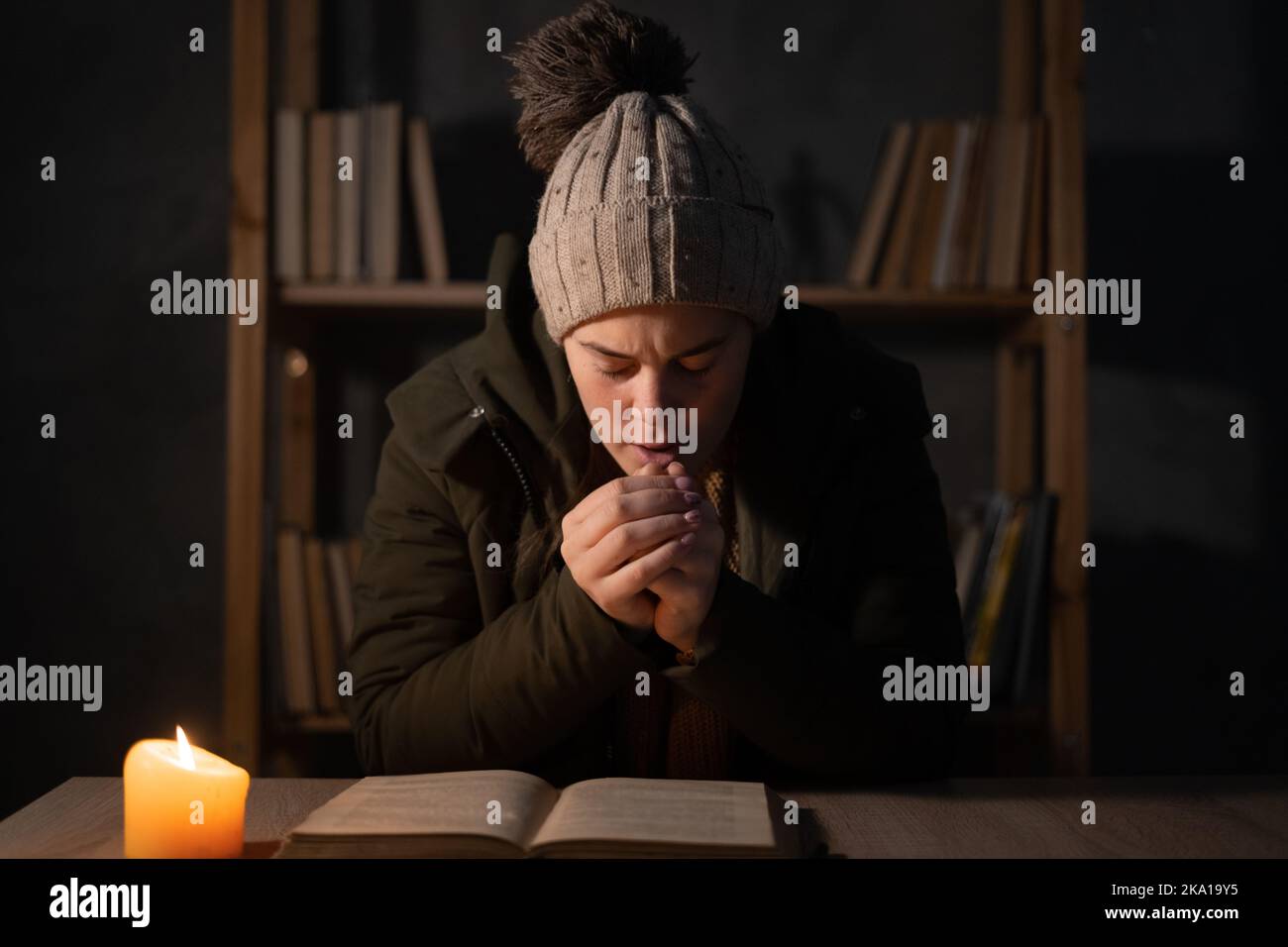 Frierende junge Frau in Winterkleidung erwärmt ihre Hände an den Lichtern mit Kerzen. Abschaltung von Heizung und Strom, Stromausfall, Stromausfall, Stromausfall, Last Stockfoto