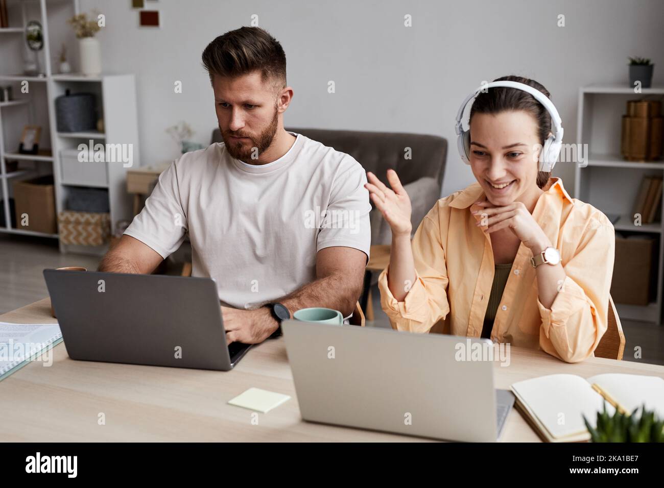 Glückliche junge Frau mit Kopfhörern, die sich im Videochat vor dem Laptop unterhielt, während sie neben einem ernsthaften bärtigen Mann am Schreibtisch saß Stockfoto