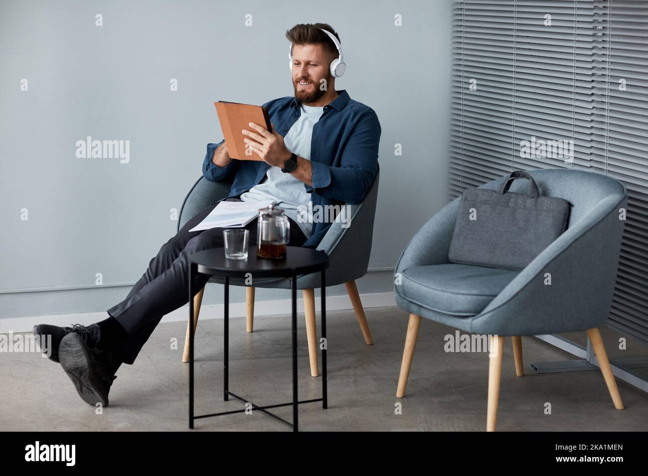 Fröhlicher junger Mann in Kopfhörern und Casualwear mit digitalem Tablet, während er im Sessel sitzt und im Videochat kommuniziert Stockfoto