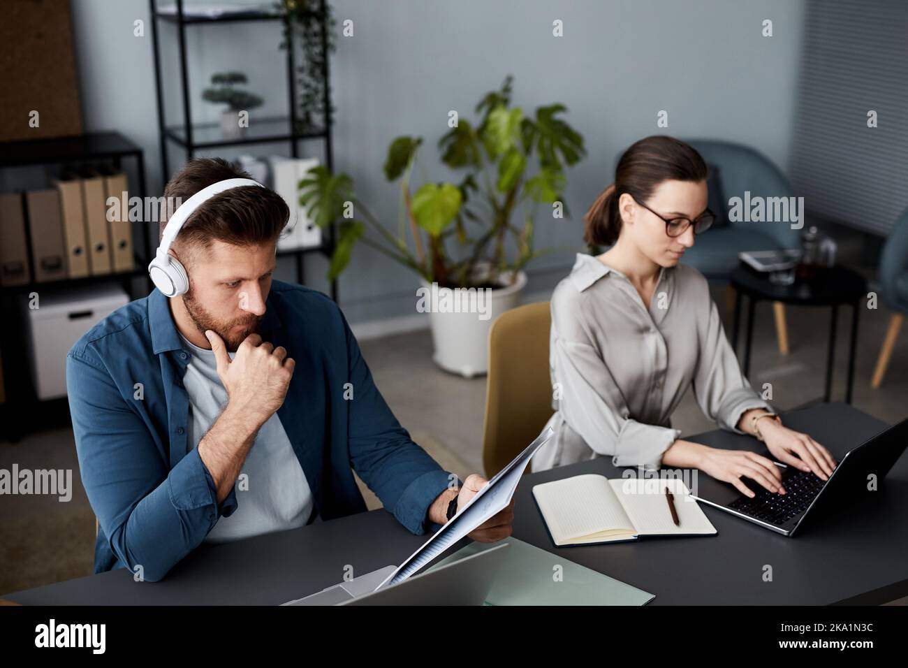Ein junger seriöser Geschäftsmann mit Kopfhörern, der Dokumente durchschaut, während sein Kollege vor dem Laptop sitzt und tippt Stockfoto