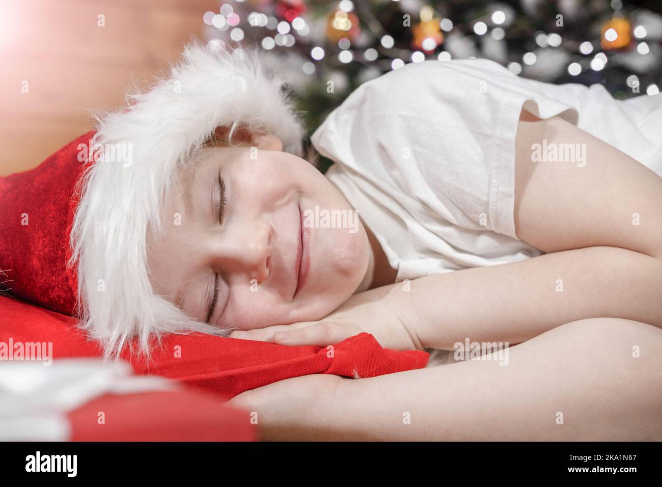 Weihnachten träumt von einem Kind. Ein Kind in einem Weihnachtsmannhut schläft im Bett neben einem Weihnachtsbaum mit Weihnachtslichtern und wartet auf das Weihnachtsgeschenk. Stockfoto