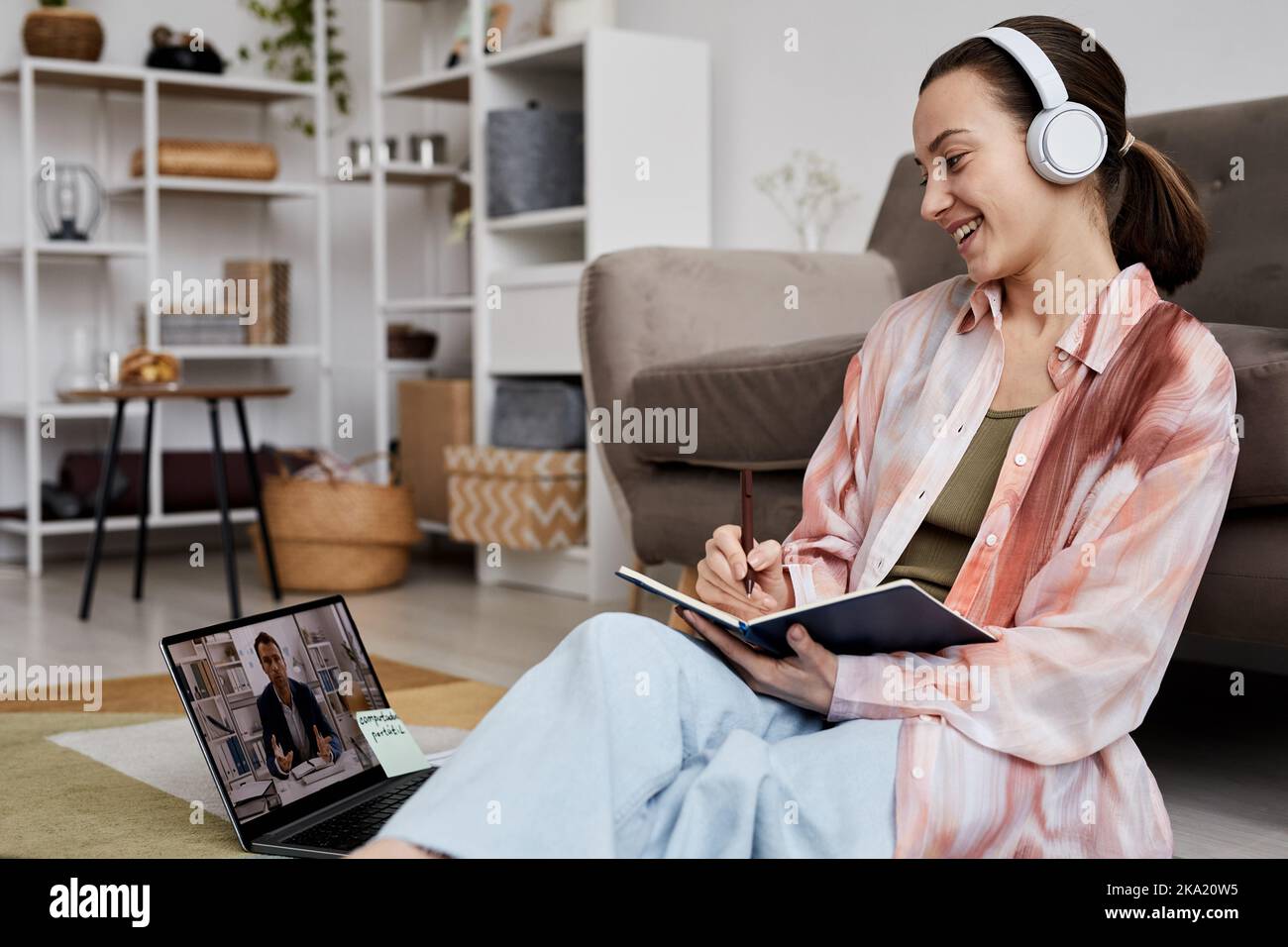 Junge, fröhliche Frau mit Kopfhörern und Casualwear, die ihrem Tutor zuhört, der während des Online-Unterrichts neues Thema erklärt Stockfoto