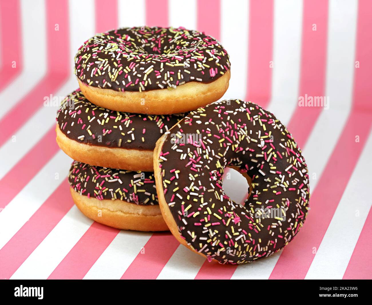 Gestapelte Donuts mit Schokoladenvereisung und farbigen Streuseln auf weiß-rosa gestreiftem Hintergrund, Nahaufnahme köstlicher Donuts Stockfoto