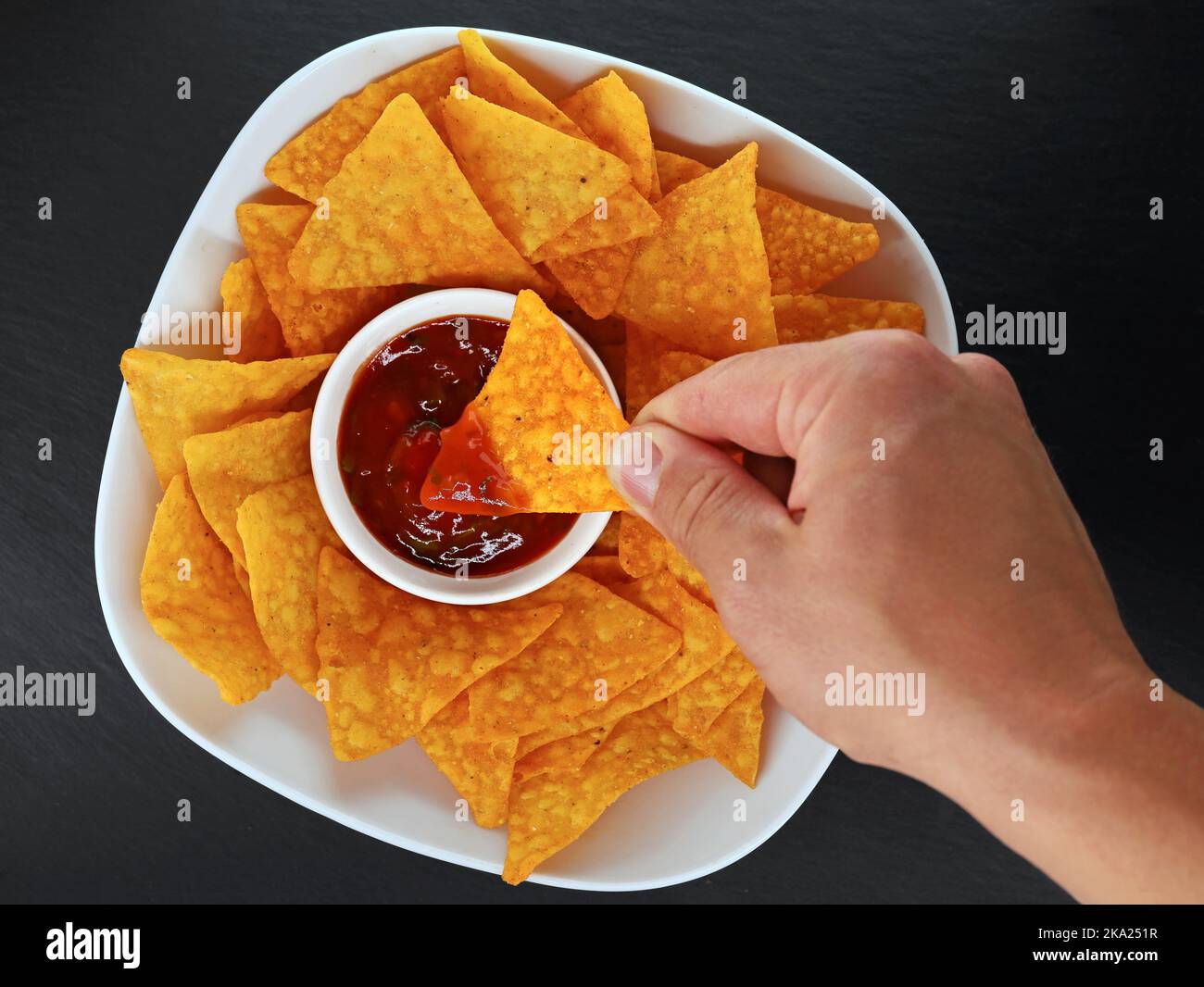 Männliche Hand taucht einen mexikanischen Maischip oder Tortilla Chip in einen würzigen Salsa Dip, Draufsicht auf eine Schüssel mit Tortillas mit Salsa auf schwarzem Schiefer Hintergrund Stockfoto