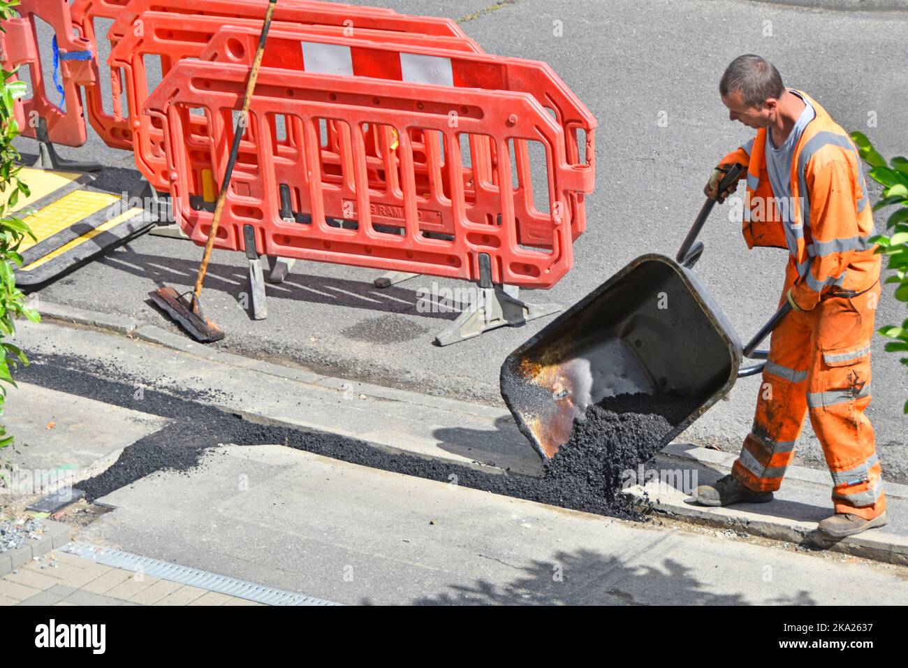 Straßenbauarbeiter mit gut sichtbarer Sicherheitskleidung, der Rückenfüllung aus der Schubkarre kippt und den Breitband-Kabelgraben auf dem Straßenbelag wiederherstellt Stockfoto